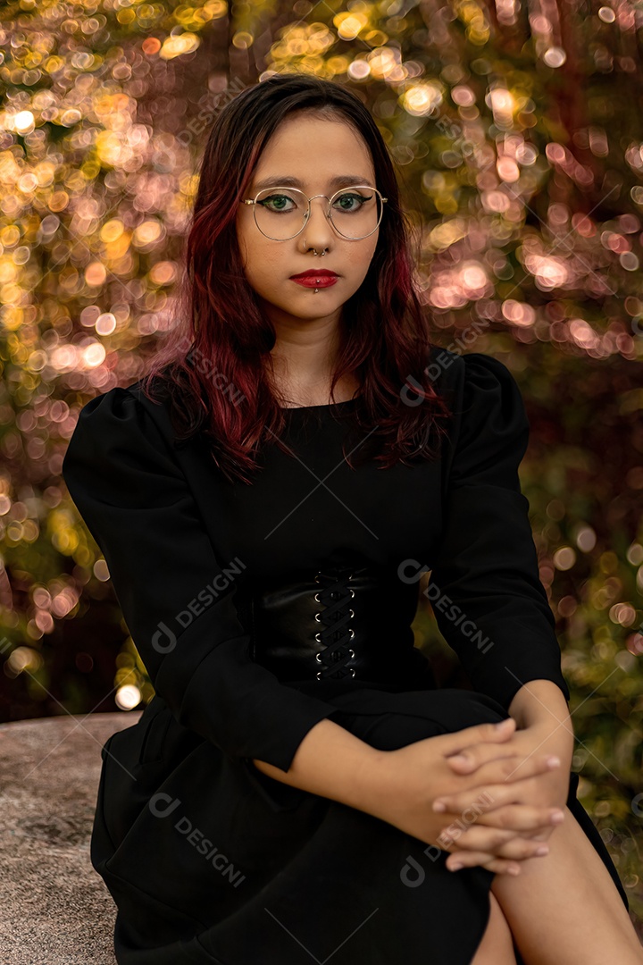 Jovem caucasiana de cabelos escuros com listras vermelhas em roupa de vestido preto com cinto espartilho e óculos posando para sessão de beleza feminina