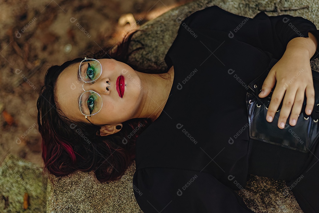 Jovem caucasiana de cabelos escuros com listras vermelhas em roupa de vestido preto com cinto espartilho e óculos posando para sessão de beleza feminina