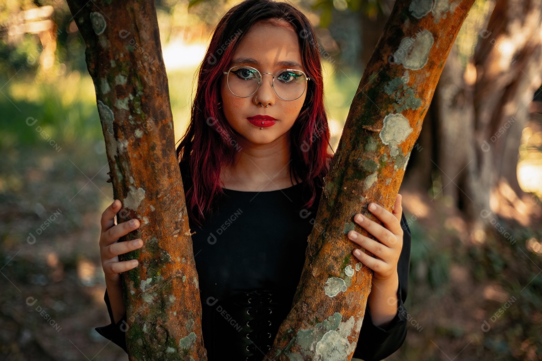 Jovem caucasiana de cabelos escuros com listras vermelhas em roupa de vestido preto com cinto espartilho e óculos posando para sessão de beleza feminina