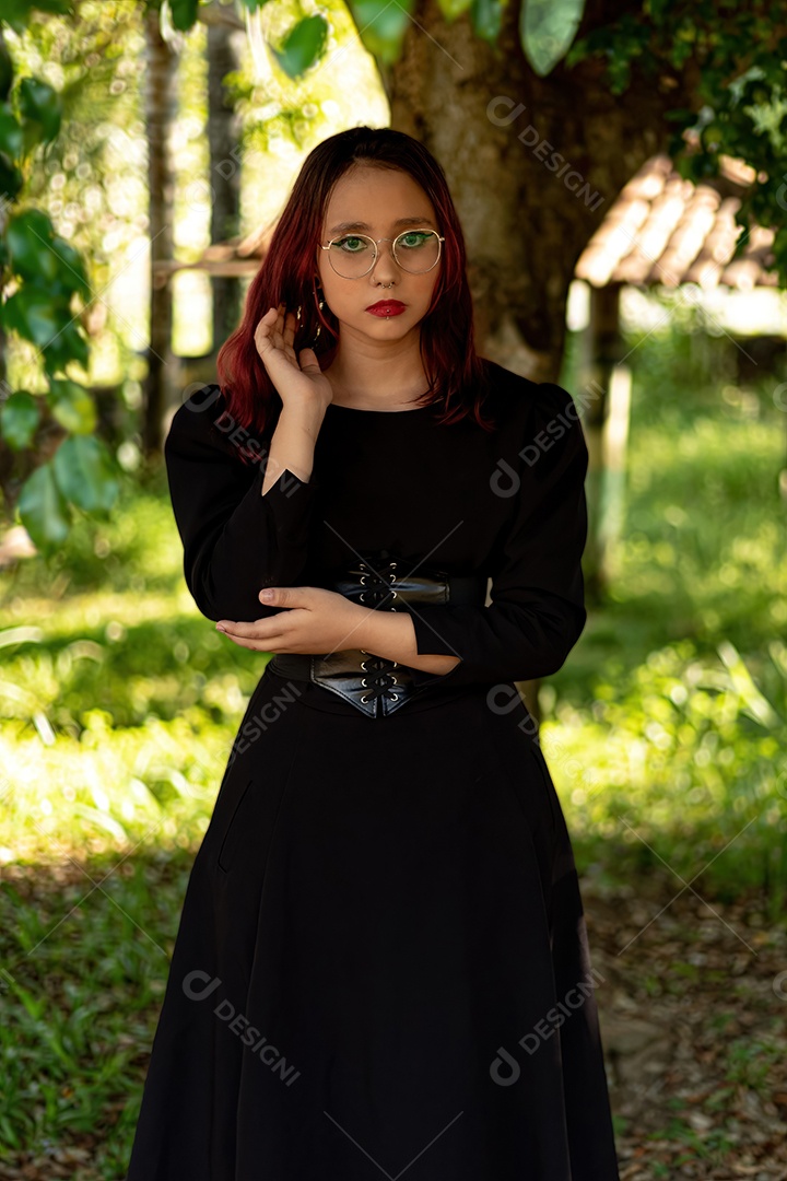 Jovem caucasiana de cabelos escuros com listras vermelhas em roupa de vestido preto com cinto espartilho e óculos posando para sessão de beleza feminina