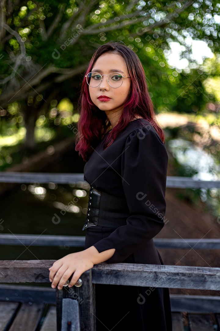 Jovem caucasiana de cabelos escuros com listras vermelhas em roupa de vestido preto com cinto espartilho e óculos posando para sessão de beleza feminina