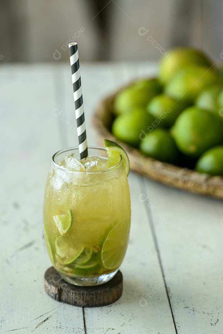 Bebida brasileira Caipirinha em uma mesa de madeira branca com limões ao fundo.