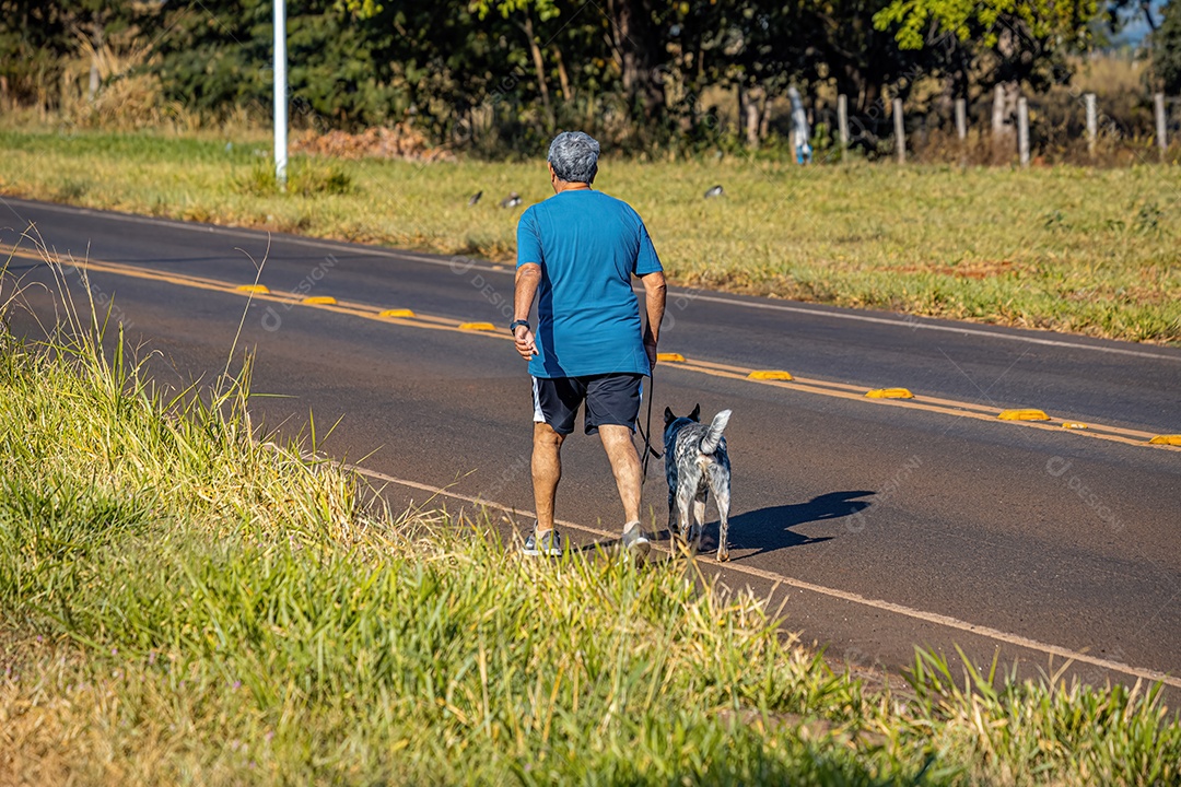 Homem caucasiano com shorts de camiseta de cabelo grisalho e sapato andando em uma estrada com seu cachorro na coleira