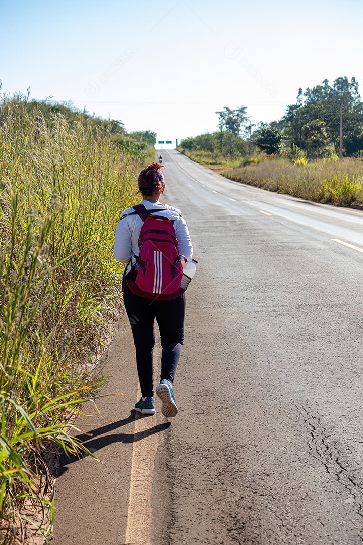 Jovem mulher com mochila rosa andando na beira de uma rodovia