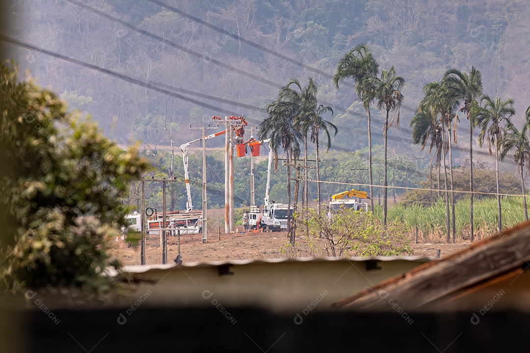 Fotografia teleobjetiva de funcionários da companhia elétrica realizando reparos em postes em uma área rural usando caminhões-guindaste.