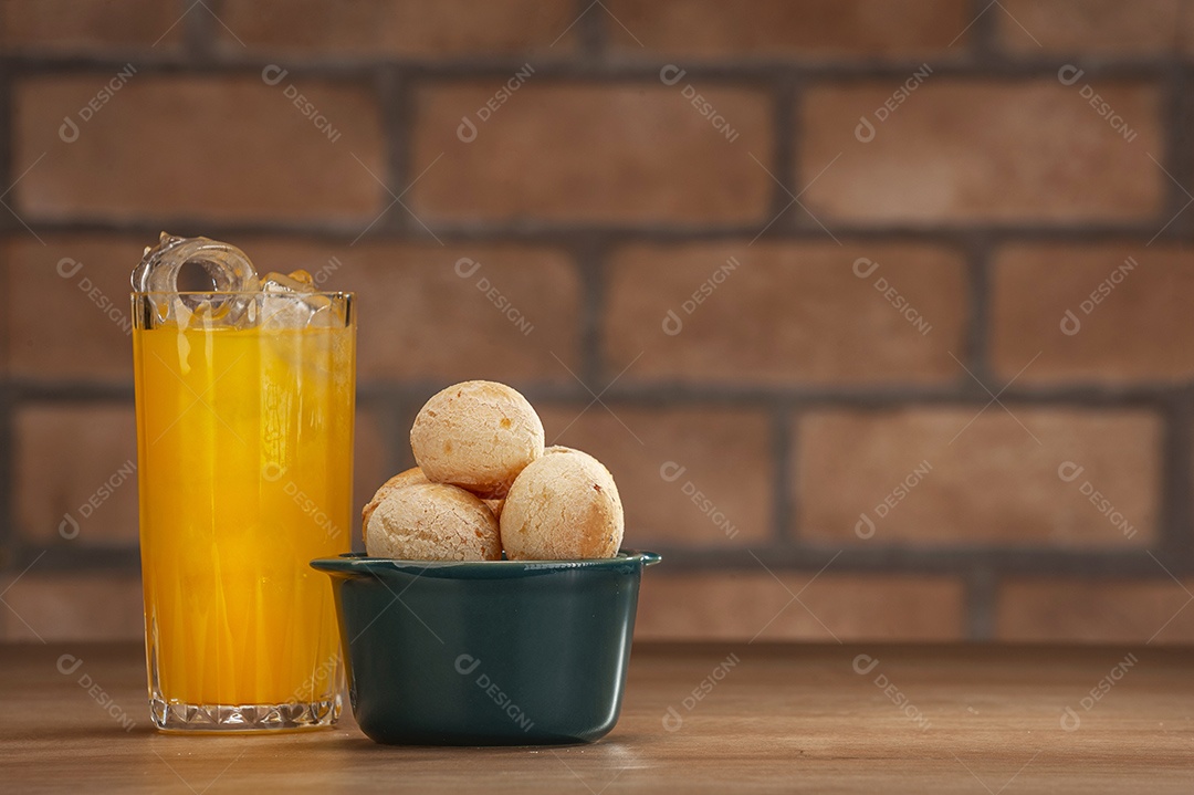 Pães de queijo em um ramekin verde com um copo de suco de laranja na mesa de madeira e fundo de parede de tijolos.