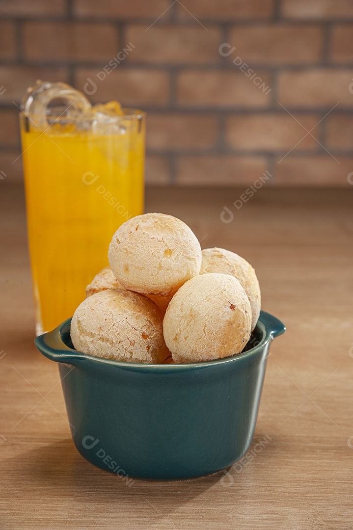 Pães de queijo em um ramekin verde com um copo de suco de laranja na mesa de madeira e fundo de parede de tijolos.
