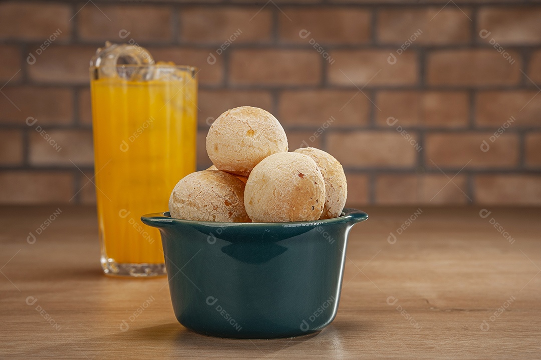Pães de queijo em um ramekin verde com um copo de suco de laranja na mesa de madeira e fundo de parede de tijolos.