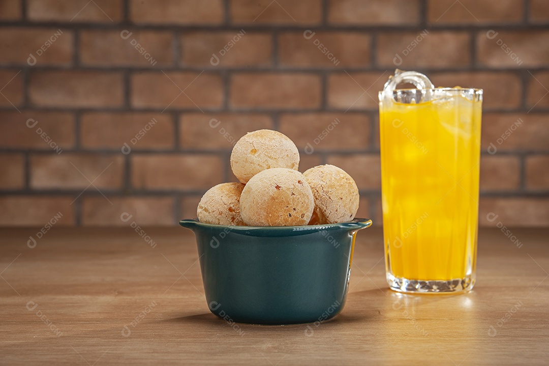 Pães de queijo em um ramekin verde com um copo de suco de laranja na mesa de madeira e fundo de parede de tijolos.