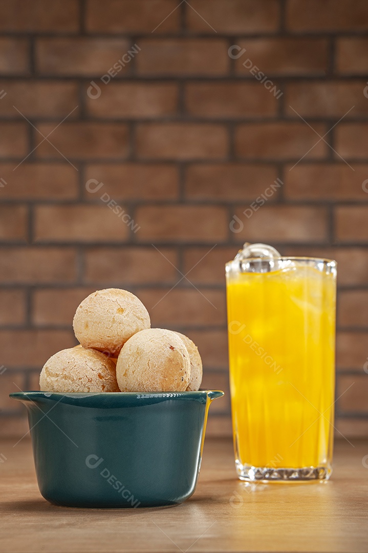 Pães de queijo em um ramekin verde com um copo de suco de laranja na mesa de madeira e fundo de parede de tijolos.