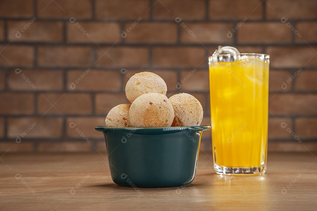 Pães de queijo em um ramekin verde com um copo de suco de laranja na mesa de madeira e fundo de parede de tijolos.