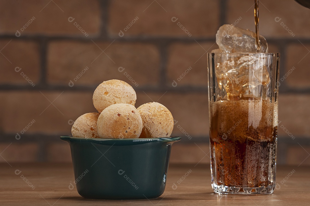 Pães de queijo em um ramekin verde com um copo de refrigerante de cola na mesa de madeira e fundo de parede de tijolos.