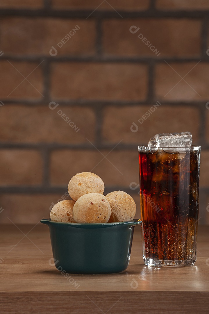 Pães de queijo em um ramekin verde com um copo de refrigerante de cola na mesa de madeira e fundo de parede de tijolos.
