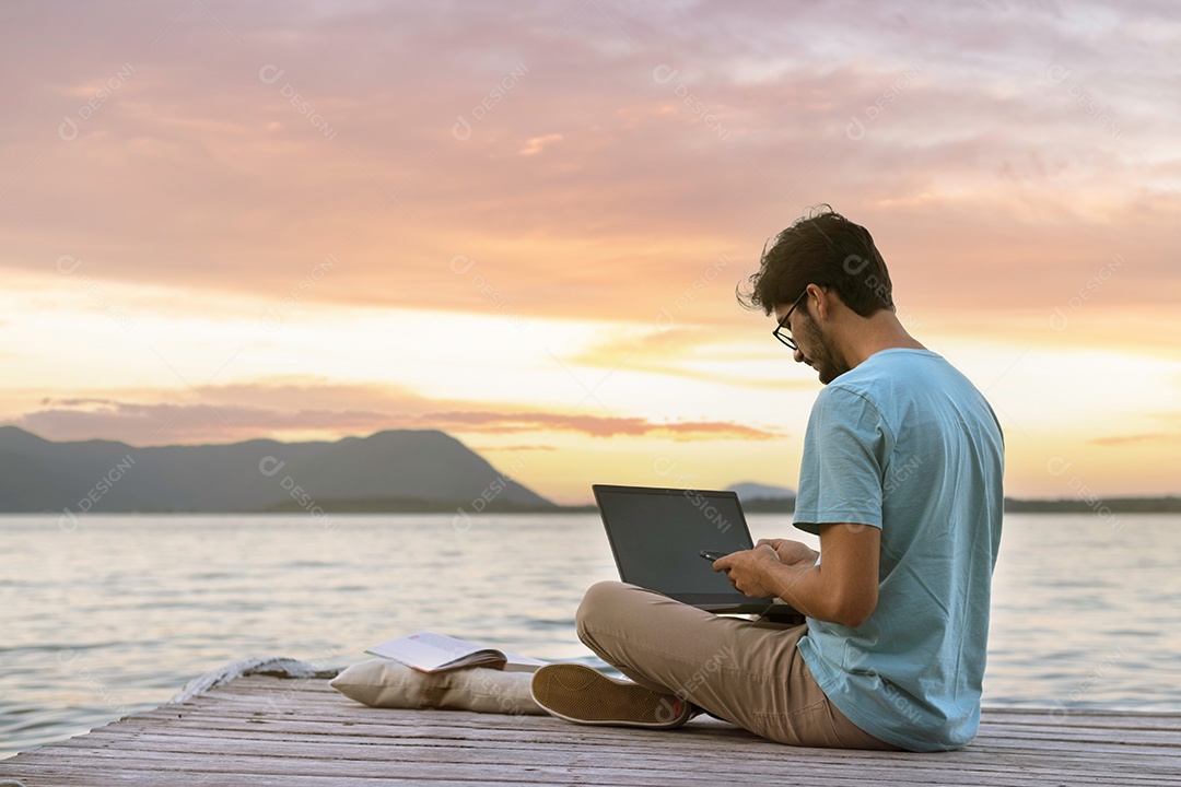 jovem nômade digital sentado no cais de madeira no mar trabalhando na internet remotamente ao pôr do sol