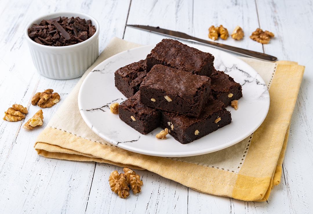 Brownies em um prato com pedaços de chocolate e nozes sobre mesa de madeira branca.