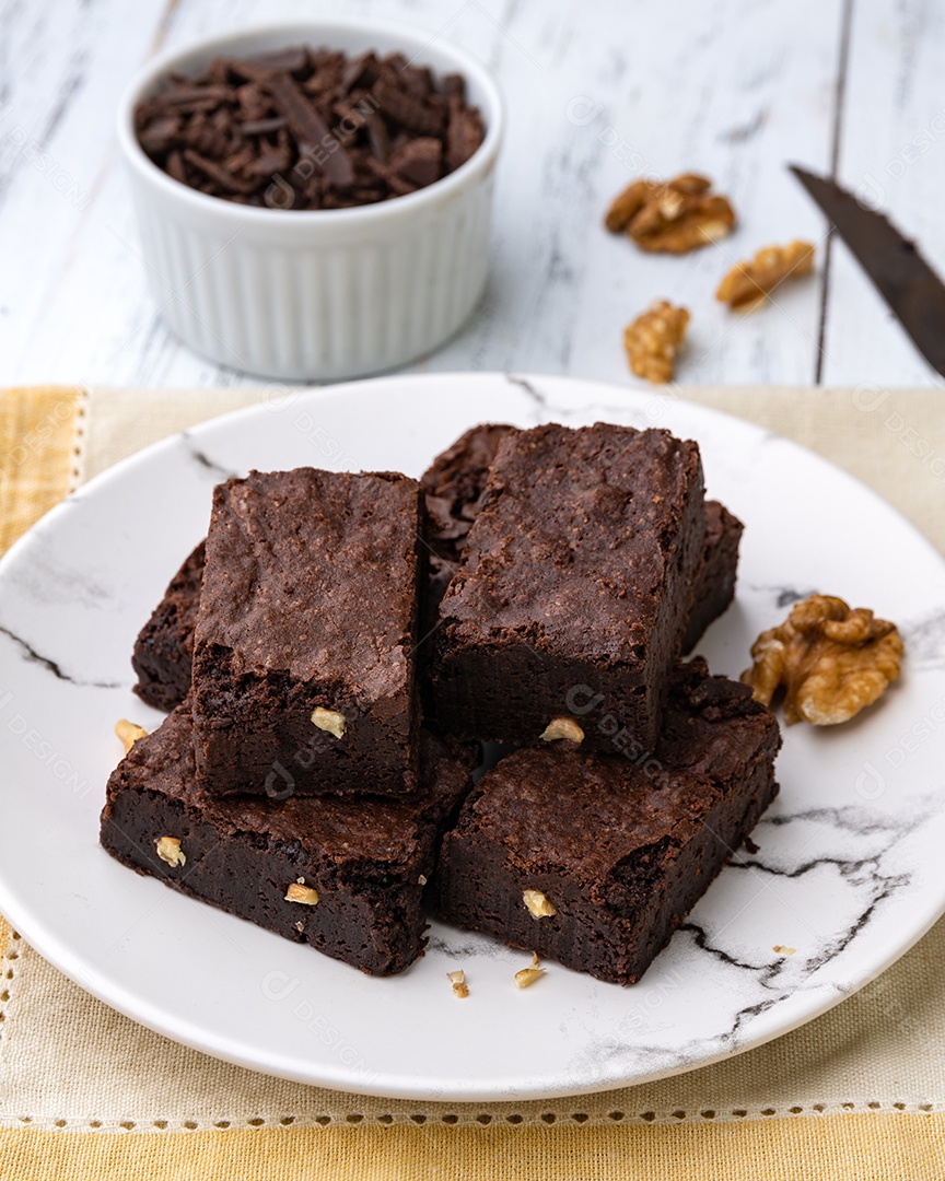 Brownies em um prato com pedaços de chocolate e nozes sobre mesa de madeira branca.