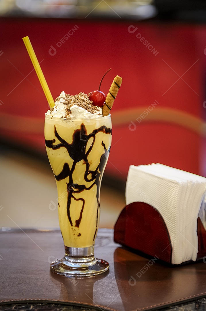 Milk-shake com calda de chocolate, chantilly e cereja, em uma mesa de café com fundo vermelho desfocado.