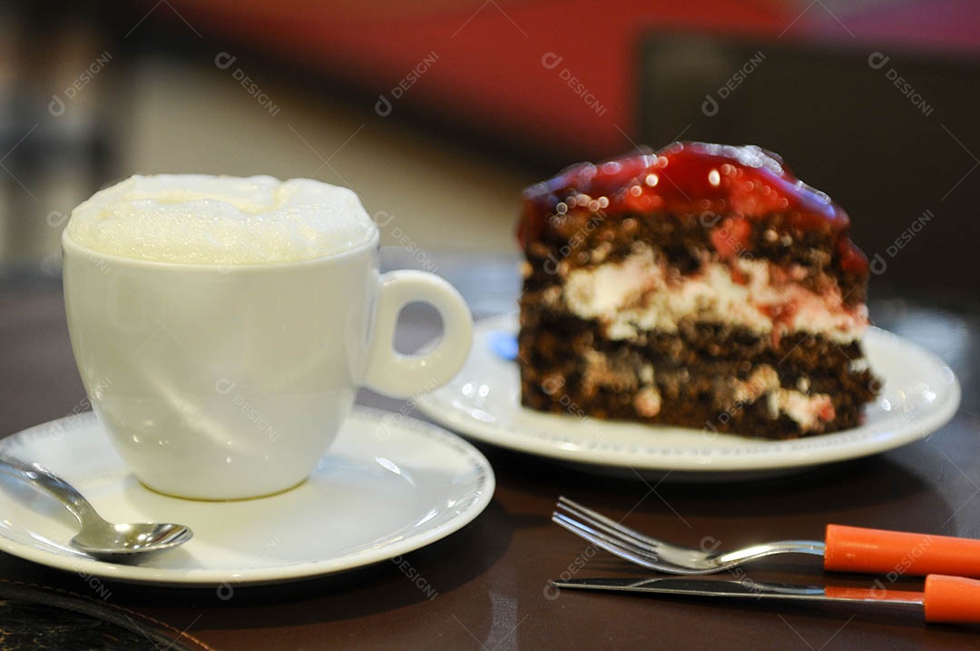 Xícara de leite quente em uma mesa de café, com uma fatia de torta de chocolate com morango no fundo desfocado.