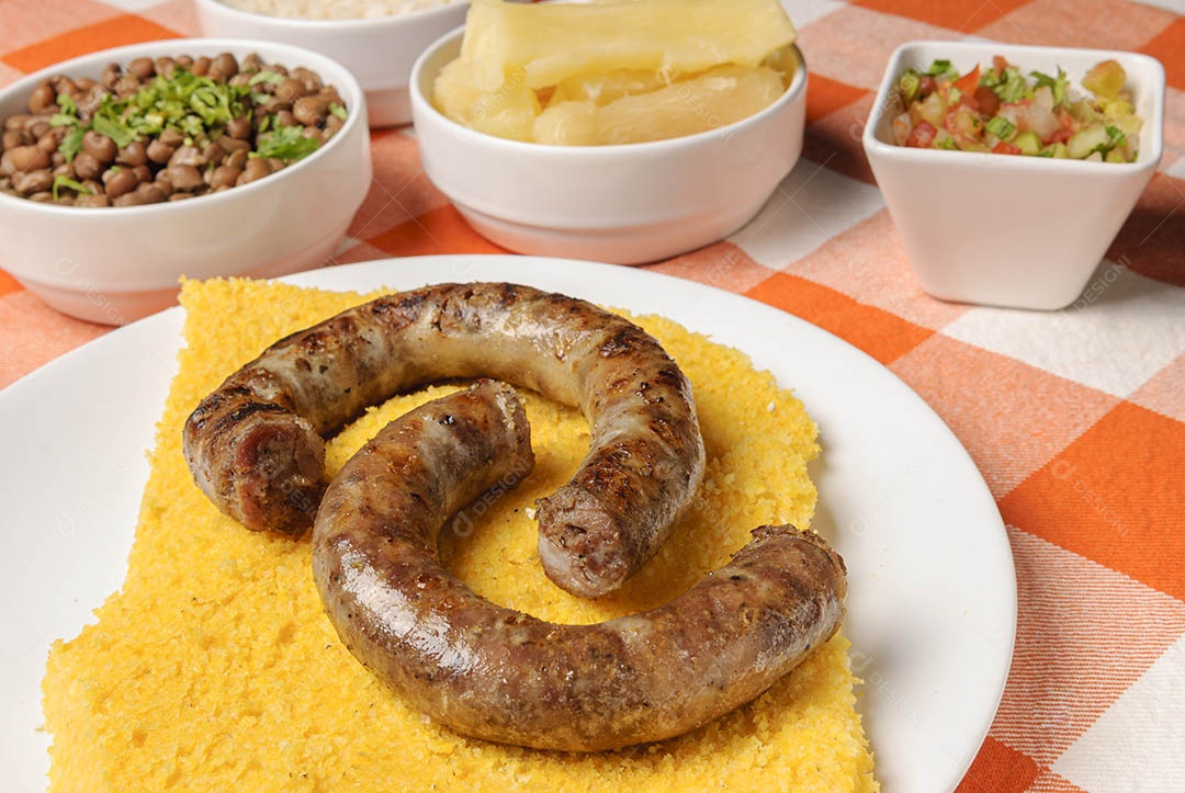 Linguiça assada com cuscuz brasileiro, acompanhada de arroz, feijão, mandioca e vinagrete, em prato branco sobre toalha quadriculada branca e laranja. Culinária nordestina brasileira.