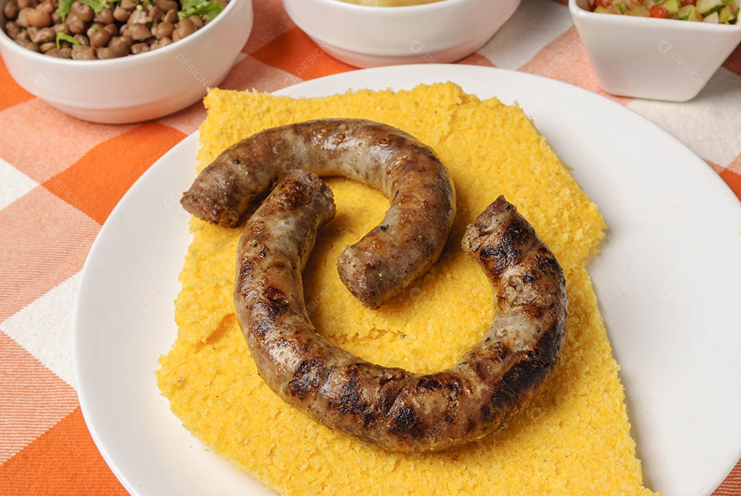 Linguiça assada com cuscuz brasileiro, acompanhada de arroz, feijão, mandioca e vinagrete, em prato branco sobre toalha quadriculada branca e laranja. Culinária nordestina brasileira.