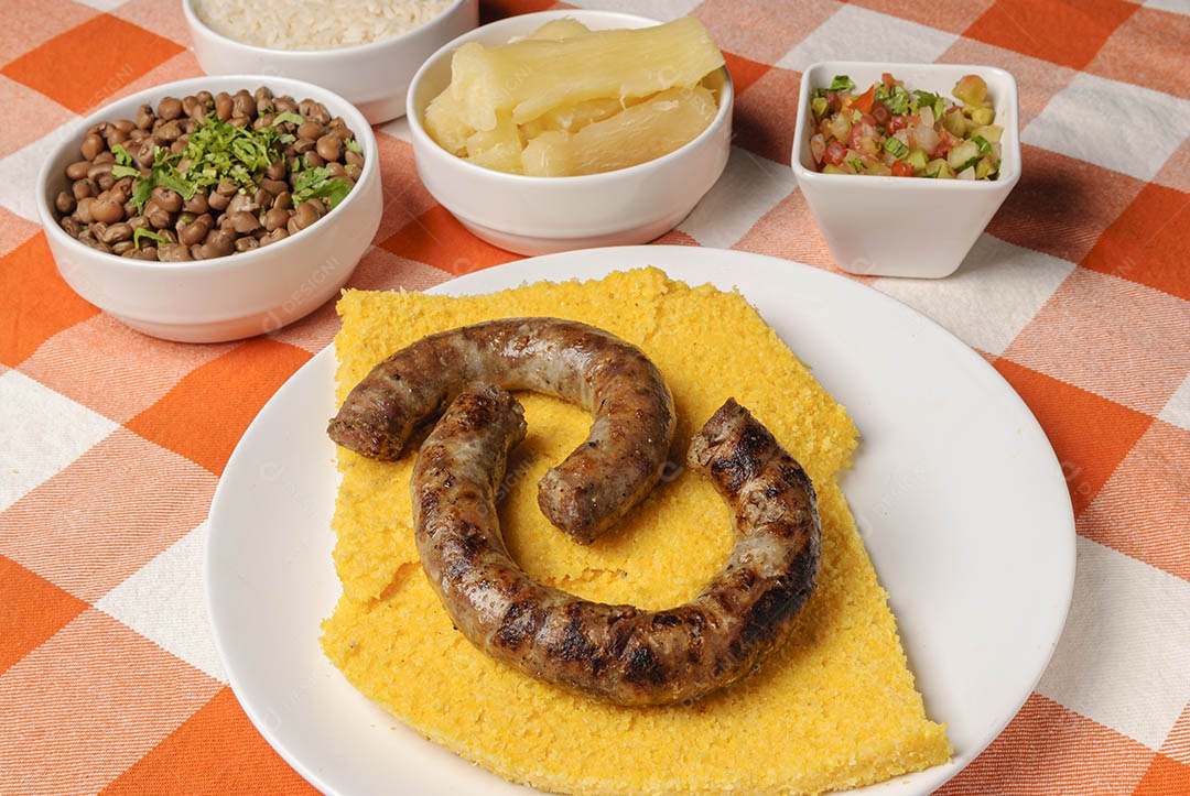 Linguiça assada com cuscuz brasileiro, acompanhada de arroz, feijão, mandioca e vinagrete, em prato branco sobre toalha quadriculada branca e laranja. Culinária nordestina brasileira.
