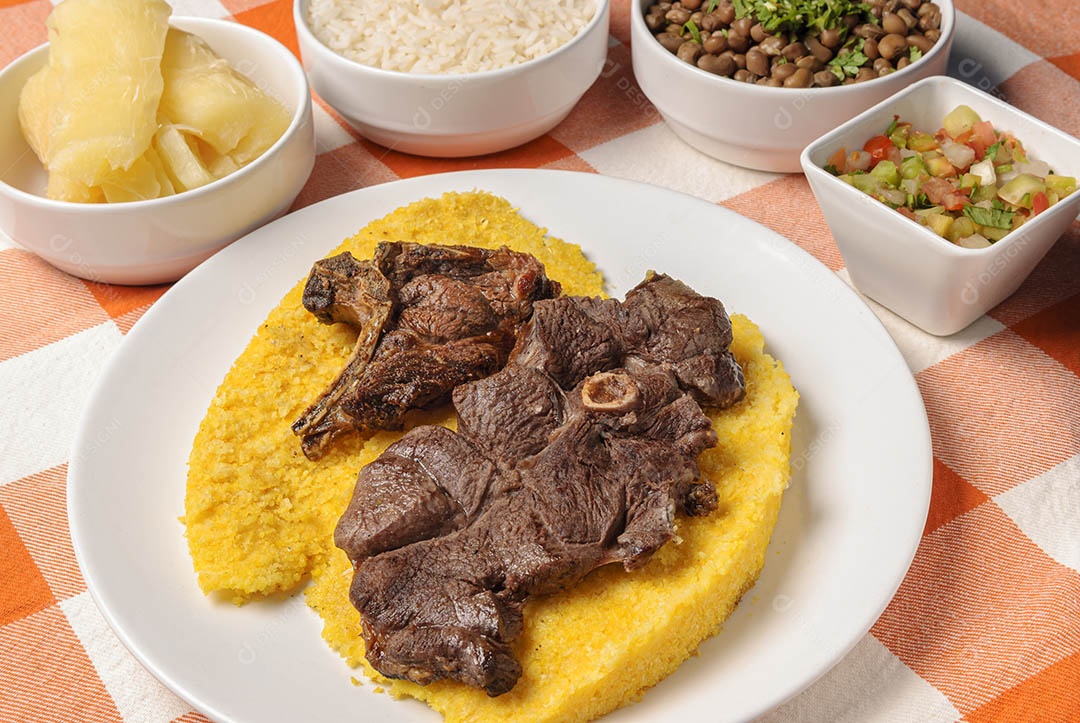 Churrasco de carne com cuscuz brasileiro, acompanhado de arroz, feijão, mandioca e vinagrete, em prato branco sobre toalha xadrez branca e laranja. Culinária nordestina brasileira.