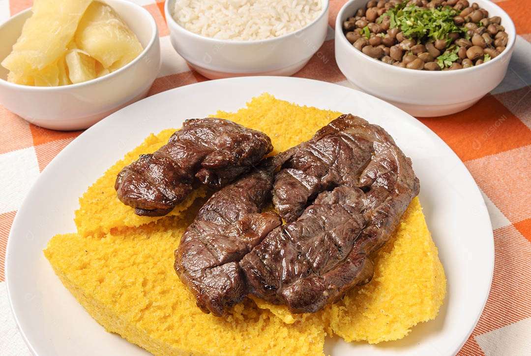 Carne assada com cuscuz brasileiro, acompanhada de arroz, feijão, mandioca e vinagrete, em prato branco sobre toalha quadriculada branca e laranja. Culinária nordestina brasileira.