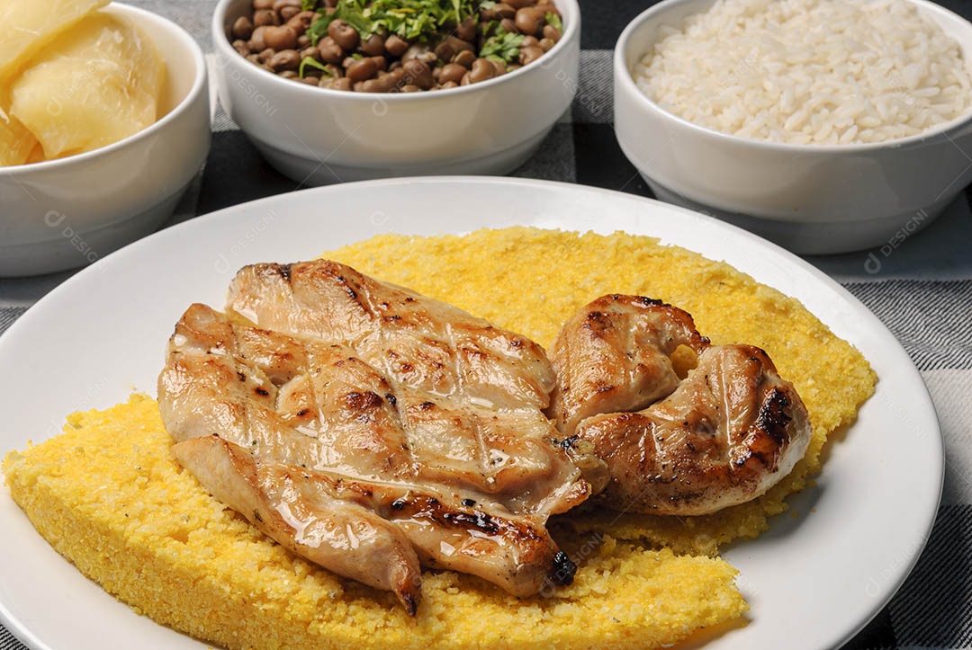 Peito de frango com cuscuz brasileiro, acompanhado de arroz, feijão, mandioca e vinagrete, em prato branco sobre toalha xadrez preto e branco. Culinária nordestina brasileira.