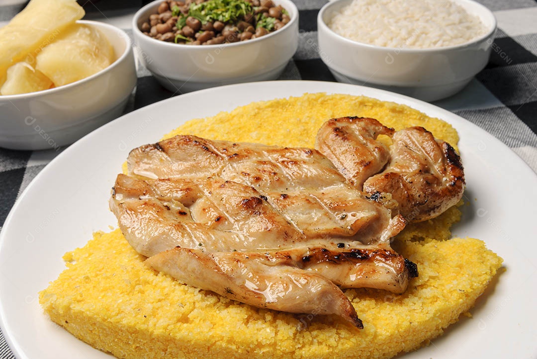 Peito de frango com cuscuz brasileiro, acompanhado de arroz, feijão, mandioca e vinagrete, em prato branco sobre toalha xadrez preto e branco. Culinária nordestina brasileira.
