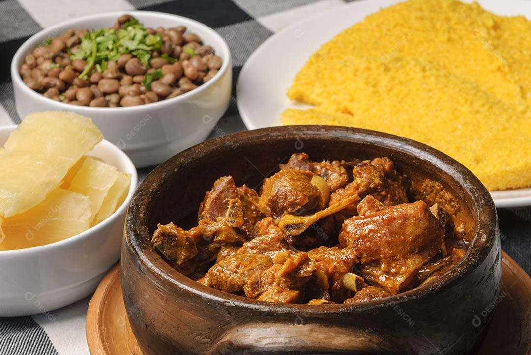 Cabrito assado com cuscuz brasileiro, acompanhado de arroz, feijão, mandioca e vinagrete, em prato branco sobre toalha quadriculada preta e branca. Culinária nordestina brasileira.