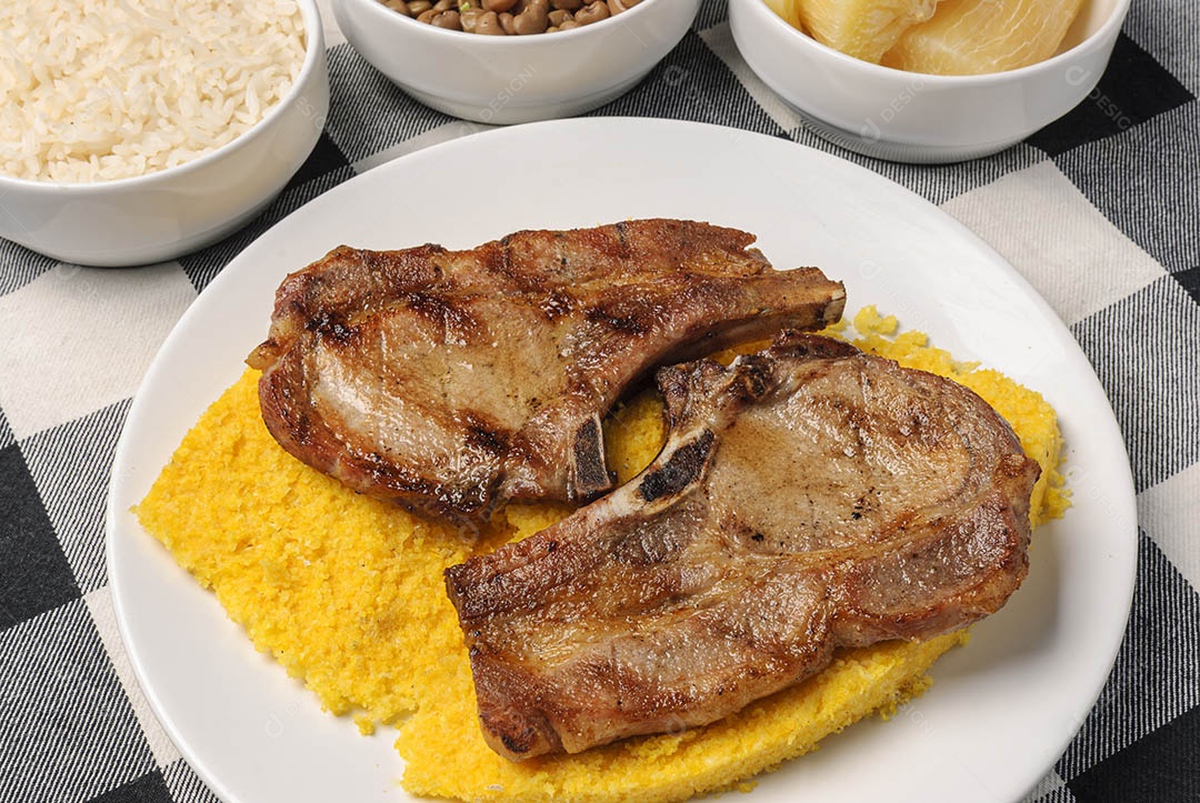 Churrasco de carne com cuscuz brasileiro, acompanhado de arroz, feijão, mandioca e vinagrete, em prato branco sobre toalha xadrez branca e laranja. Culinária nordestina brasileira.