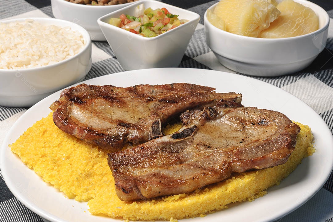 Churrasco de carne com cuscuz brasileiro, acompanhado de arroz, feijão, mandioca e vinagrete, em prato branco sobre toalha xadrez branca e laranja. Culinária nordestina brasileira.