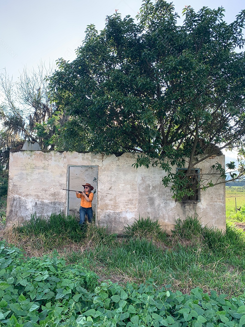 Mulher loira brasileira segurando uma espingarda em uma casa abandonada no brasil. Camisa laranja e chapéu.