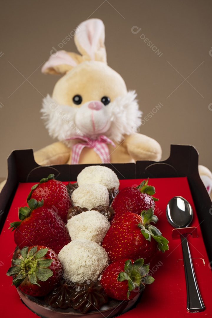 Caixa vermelha com meio ovo de páscoa de chocolate e um coelhinho de pelúcia ao fundo.