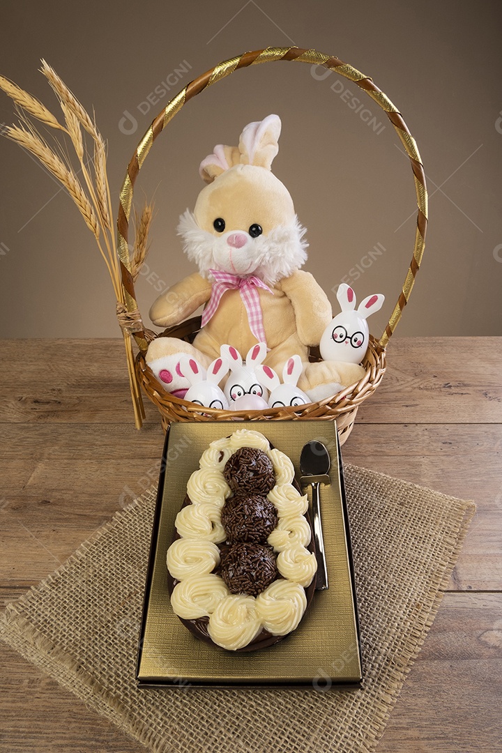 Caixa dourada com meio ovo de páscoa e um coelhinho de pelúcia ao fundo.