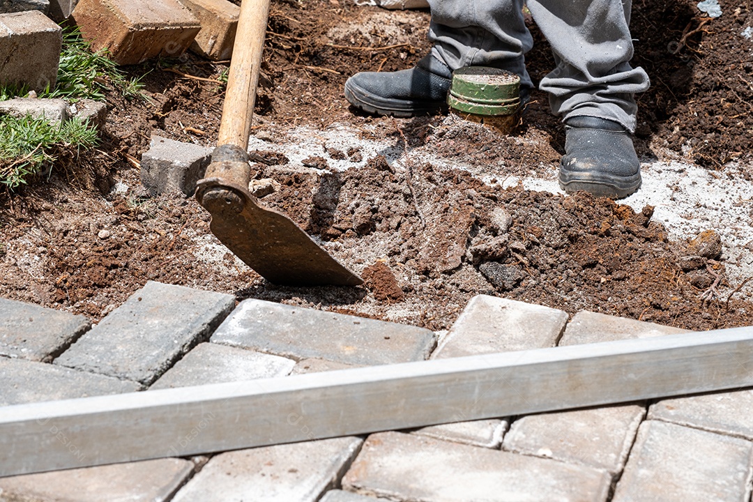 Brasileiro usando enxada para preparar o solo para o assentamento de blocos de concreto.
