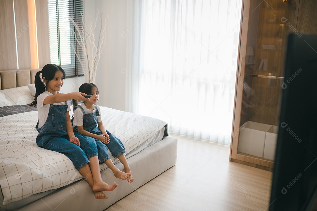 Meninas asiáticas irmãs com controle remoto sentadas na cama assistindo a um filme na tv