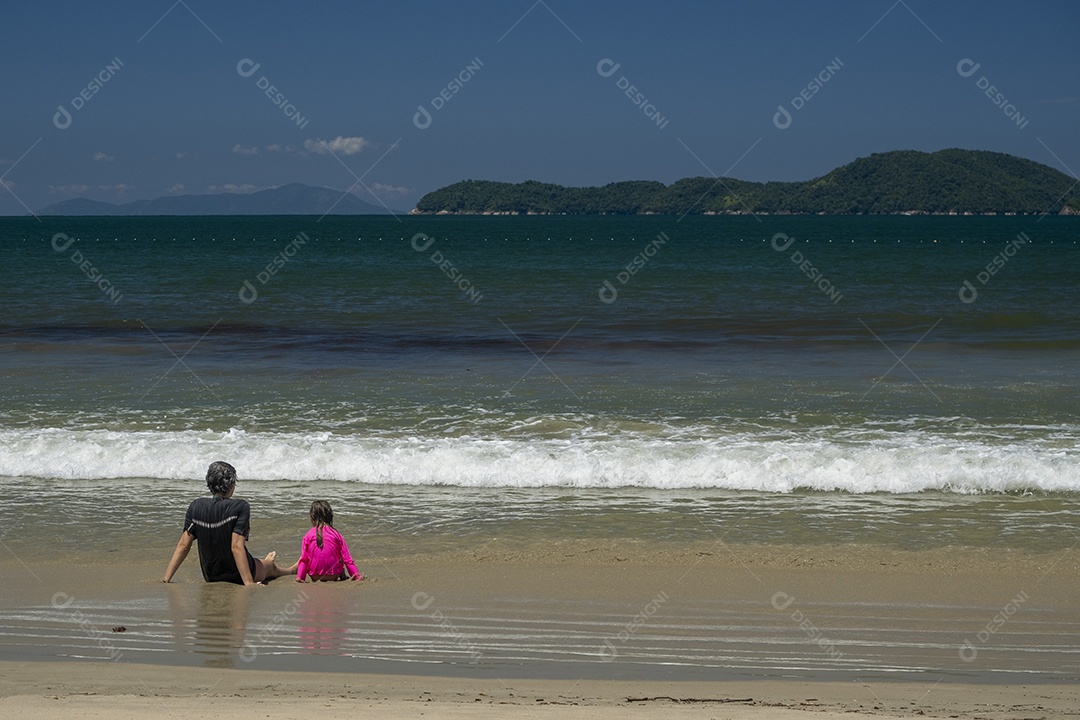 Mãe e filha sentadas na areia da praia olhando o mar