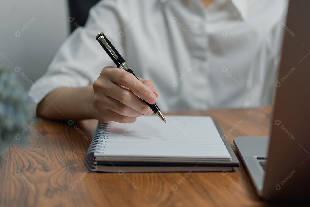 mão de uma mulher segurando uma caneta e escrevendo informações em um caderno trabalhando com computador portátil.