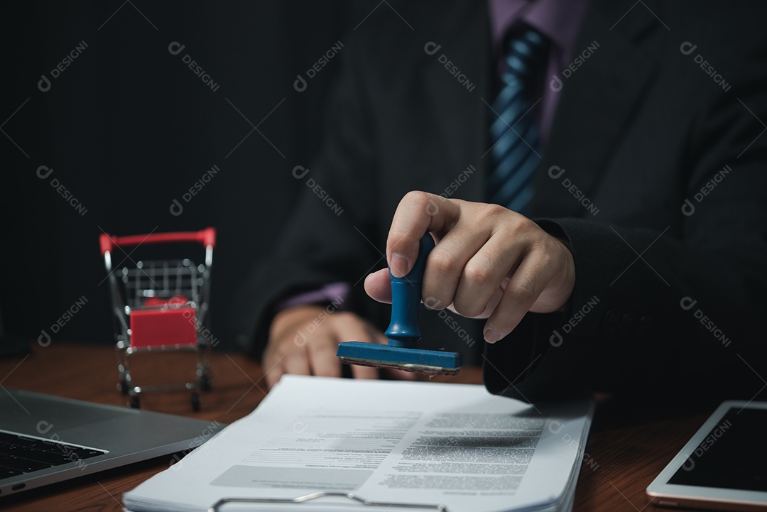 Mão de homem carimbando aprovação assinando contrato de documento ou papelada na mesa.