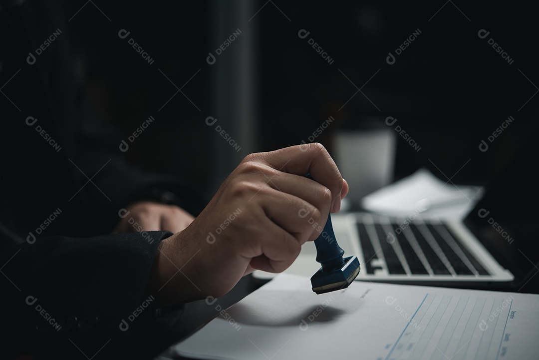 Mão de homem carimbando aprovação assinando contrato de documento ou papelada na mesa.