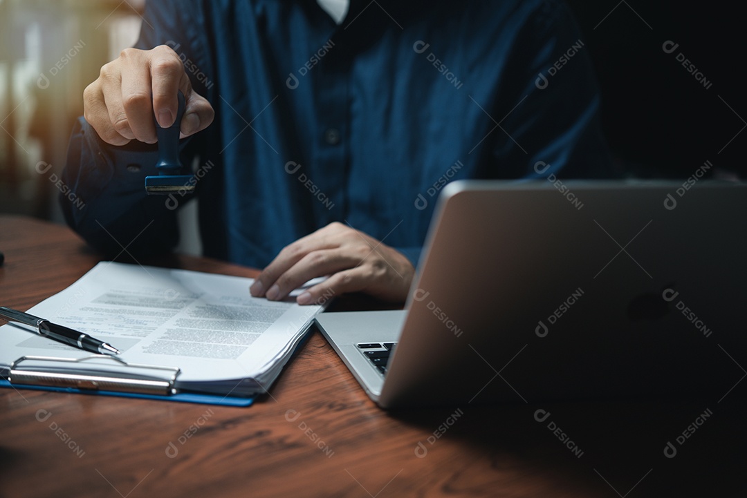 Mão de homem carimbando aprovação assinando contrato de documento ou papelada na mesa.