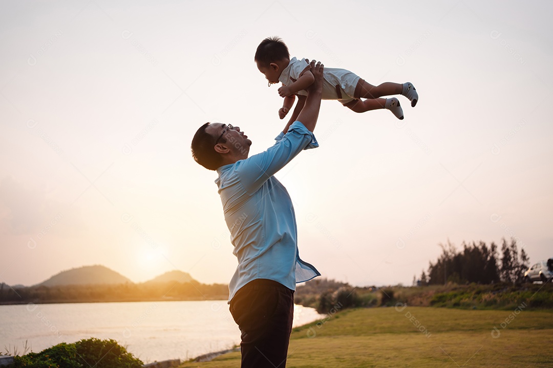 Paternidade levantando filho bebê desfrutando no parque perto do rio ao pôr do sol.