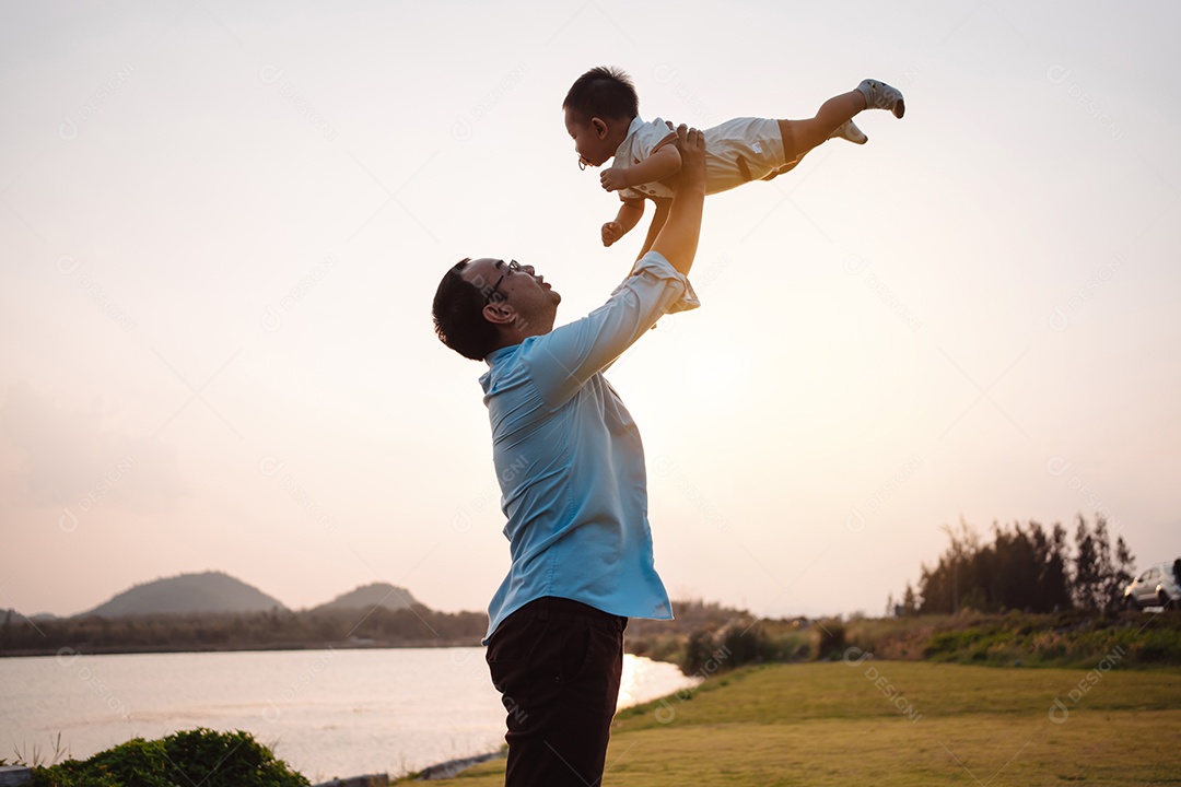 Paternidade levantando filho bebê desfrutando no parque perto do rio ao pôr do sol.