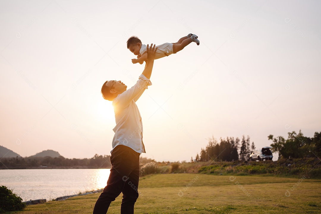 Paternidade levantando filho bebê desfrutando no parque perto do rio ao pôr do sol.