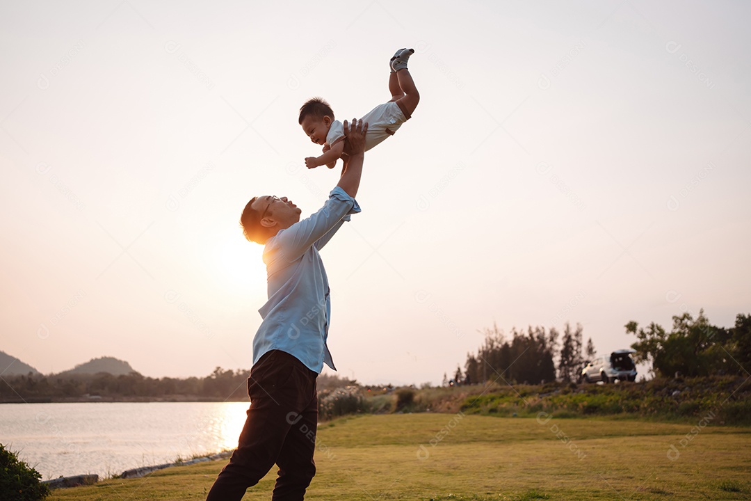 Paternidade levantando filho bebê desfrutando no parque perto do rio ao pôr do sol.