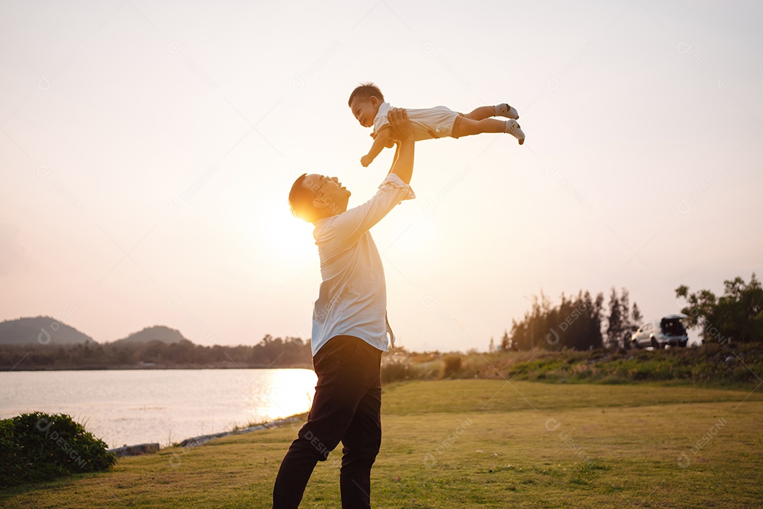 Paternidade levantando filho bebê desfrutando no parque perto do rio ao pôr do sol.