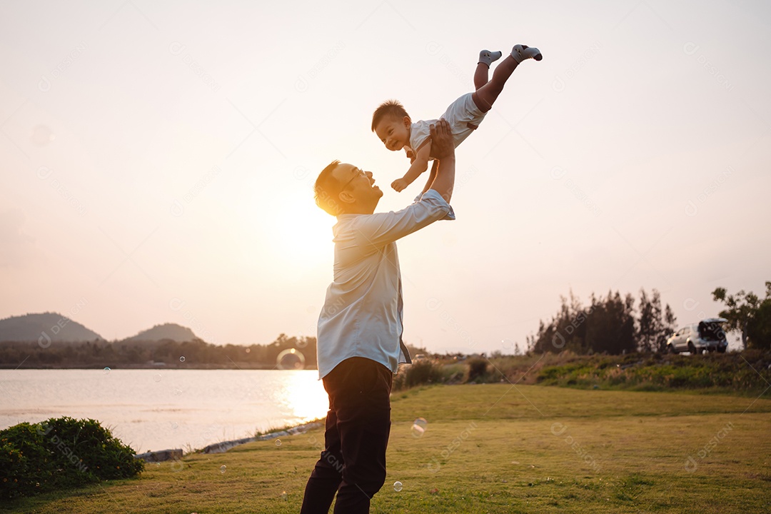 Paternidade levantando filho bebê desfrutando no parque perto do rio ao pôr do sol.