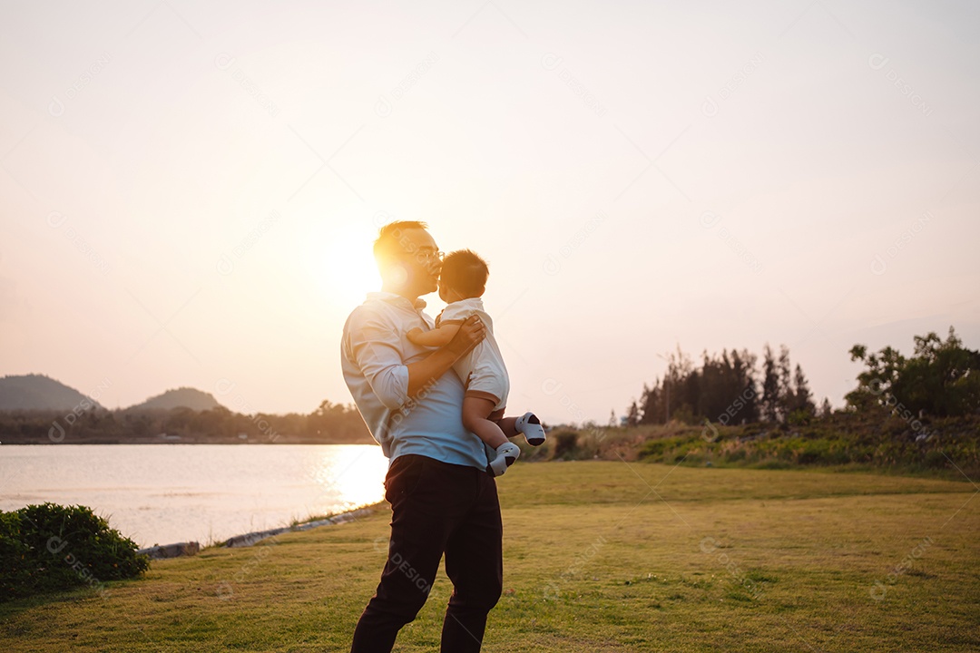 Paternidade levantando filho bebê desfrutando no parque perto do rio ao pôr do sol.