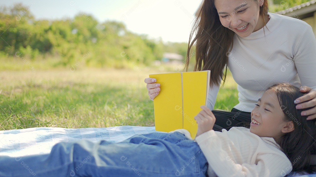 Bela jovem asiática mãe e filha fazendo piquenique e lendo conto em livros amarelos no parque verde.
